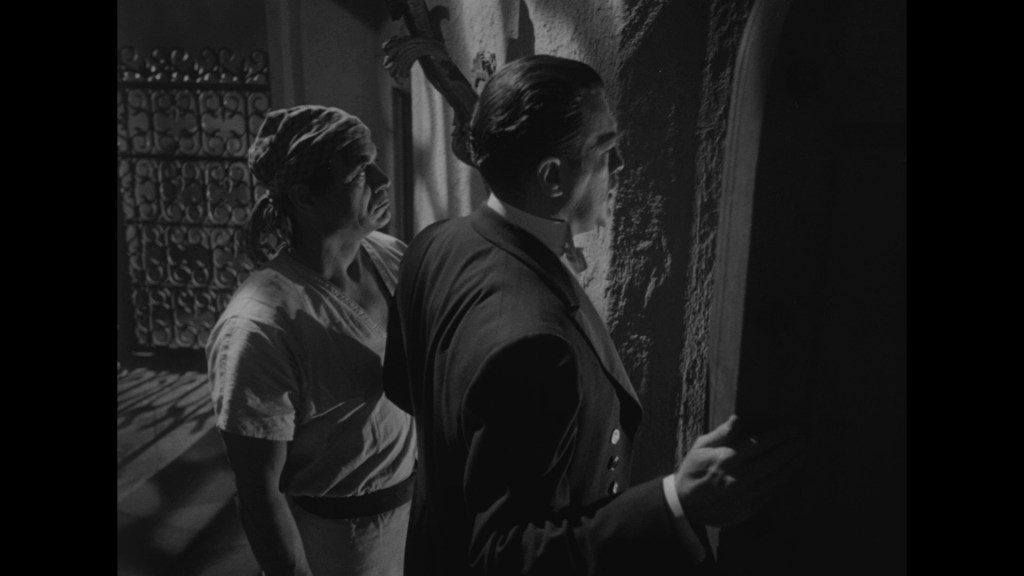 Two men stand in a dimly lit setting. One man, wearing a formal suit, is looking at a door while the other, dressed in a simple shirt, gazes at him with a concerned expression.