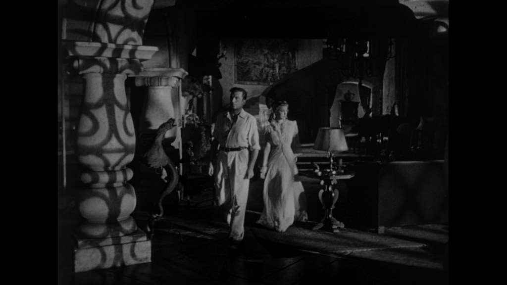 A man and a woman walking through a dimly lit room with ornate decor and shadows cast by furniture.
