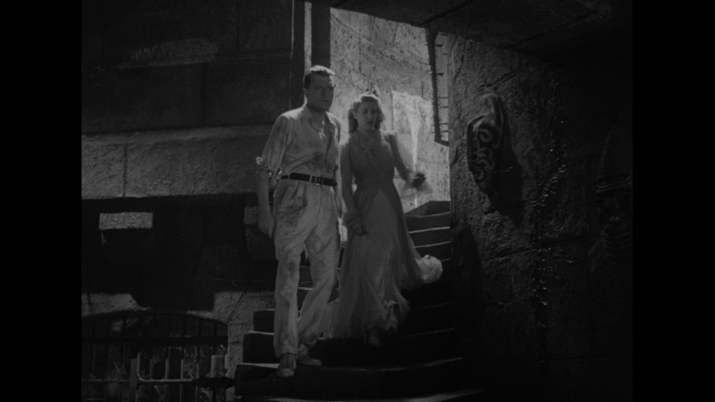 A man and a woman walking down a stone staircase in a dimly lit environment, with ancient carvings on the walls.