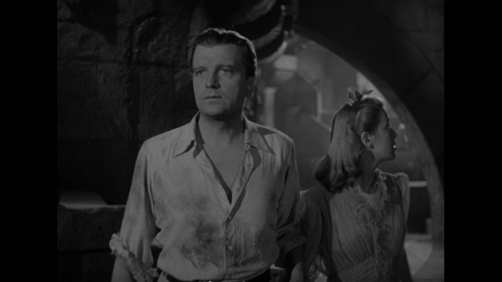 A man and a woman stand together in a dimly lit, atmospheric setting, looking off-screen with expressions of concern or fear.