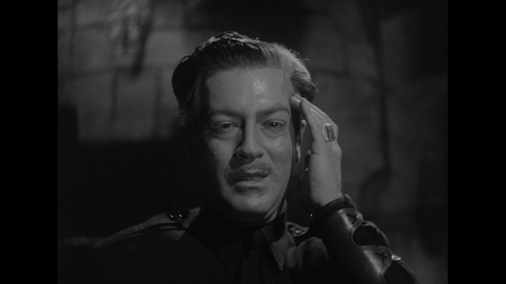 A dramatic black and white portrait of a man with slicked-back hair and a mustache, wearing a uniform. He has a pained expression, touching his forehead with one hand and showing a ring on his other hand.