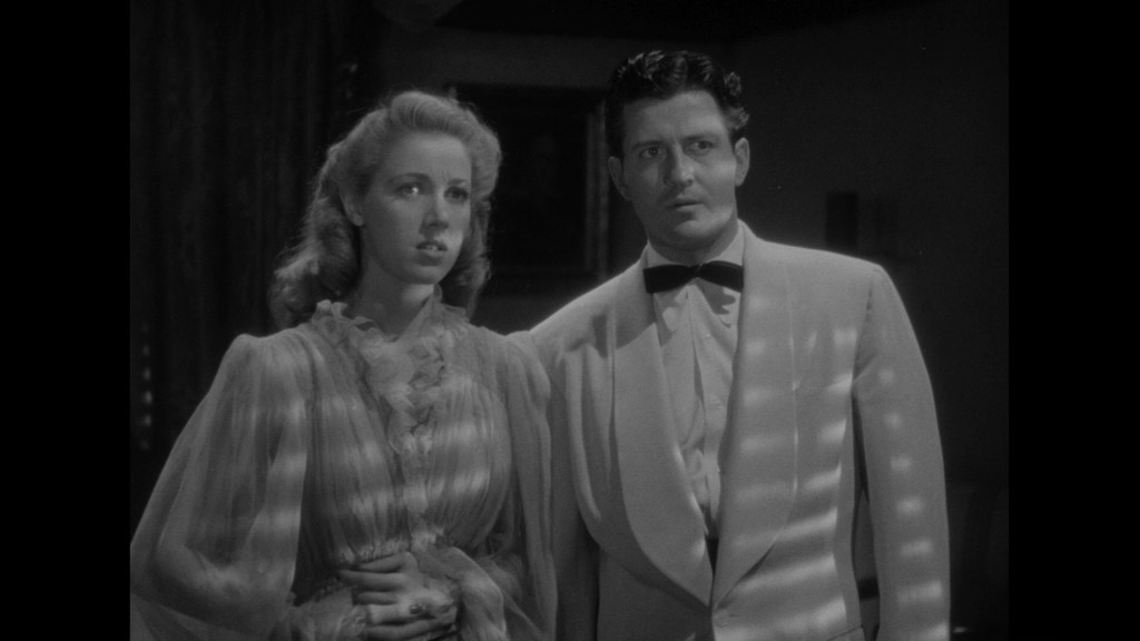 A shocked man in a tuxedo and a woman in a flowing dress stand together, with light casting striped shadows across them, creating a dramatic atmosphere.