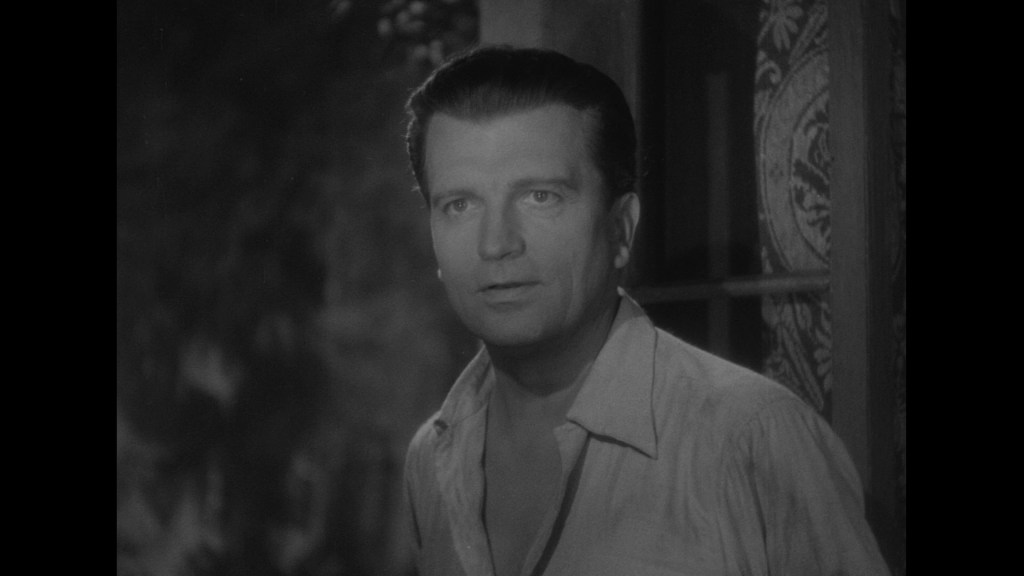 A man with slicked-back hair and a concerned expression, standing in front of a window with patterned curtains, captured in black and white.