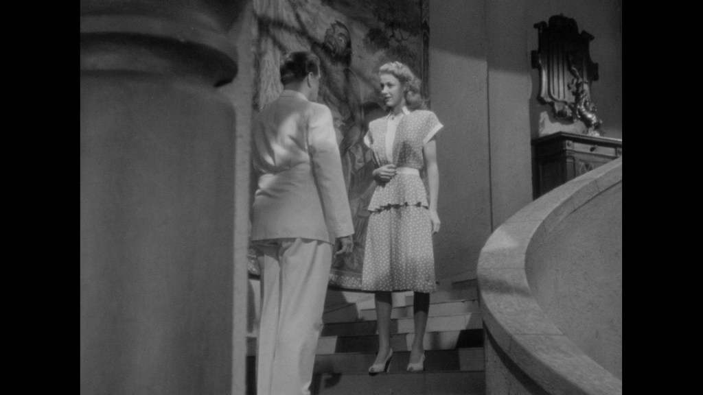 A man in a white suit stands at the bottom of a staircase, looking up at a woman dressed in a polka dot dress, who is on the stairs. The scene is in black and white with a decorative background.