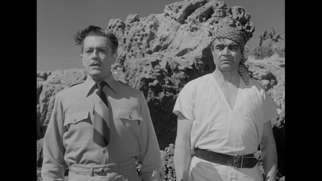Two men standing in front of rocky terrain, one in military uniform and the other in a simple white garment with a headscarf.