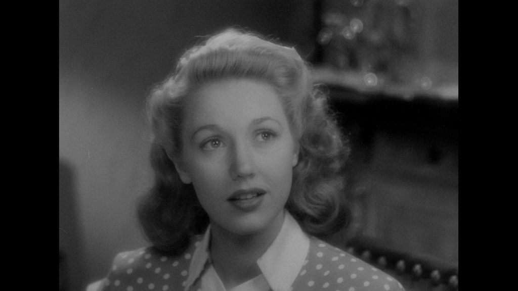 Close-up portrait of a woman with wavy hair, wearing a polka dot blouse, looking thoughtfully with a soft expression in a vintage black and white setting.