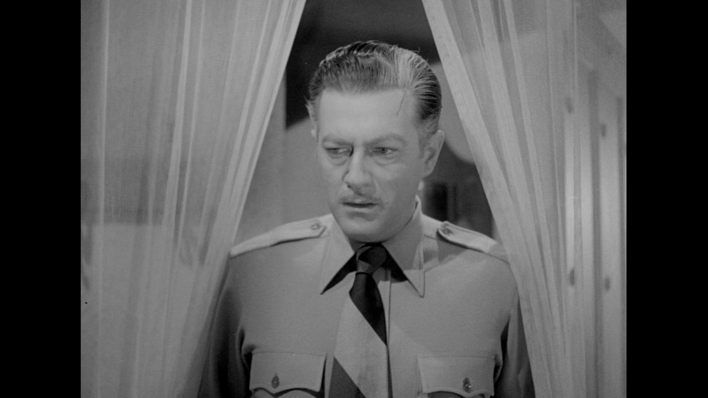A man wearing a military uniform stands behind sheer curtains, looking intently to the side.