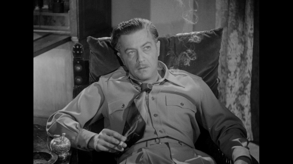 A man wearing a military-style outfit with a cigarette, sitting in a chair with a contemplative expression, smoke rising around him.