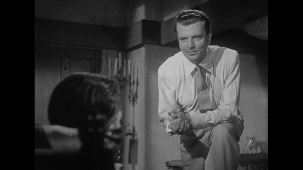 A man in a white shirt and tie leans forward, engaging with another person off-camera in a dimly lit room with candles and rustic decor.