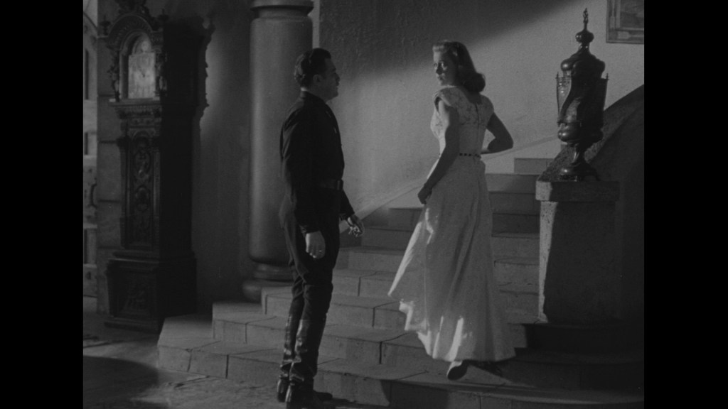 A man wearing a dark uniform stands at the bottom of a staircase, looking up at a woman in a white dress as she ascends the stairs.