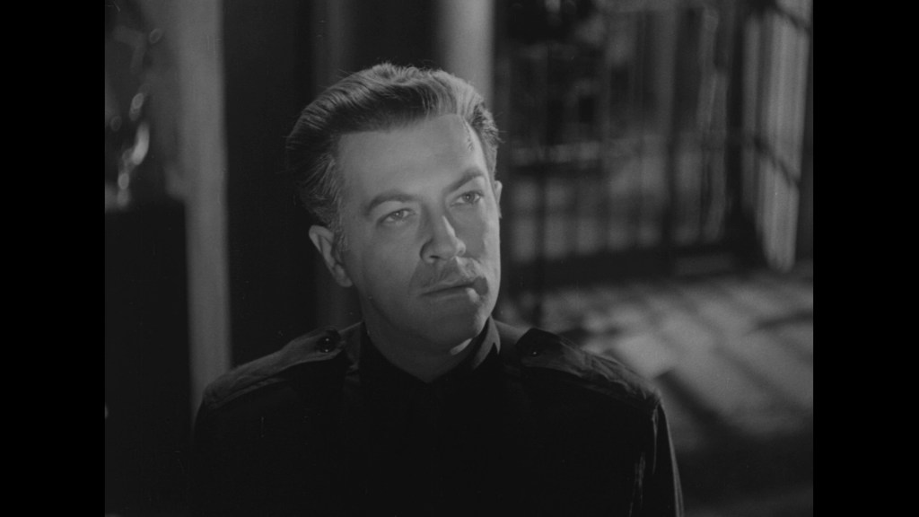 A close-up of a man with slicked-back hair and a mustache, wearing a dark uniform, looking contemplatively to the side in a black and white scene.