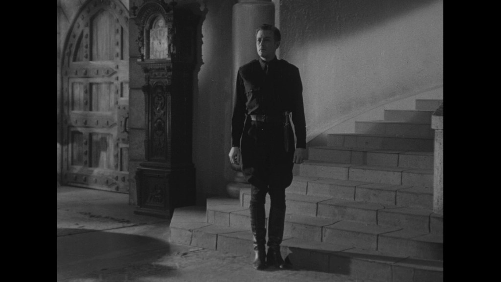 A man in a dark outfit stands on a stairway in a dimly lit interior, with an ornate wooden door and a decorative structure in the background.