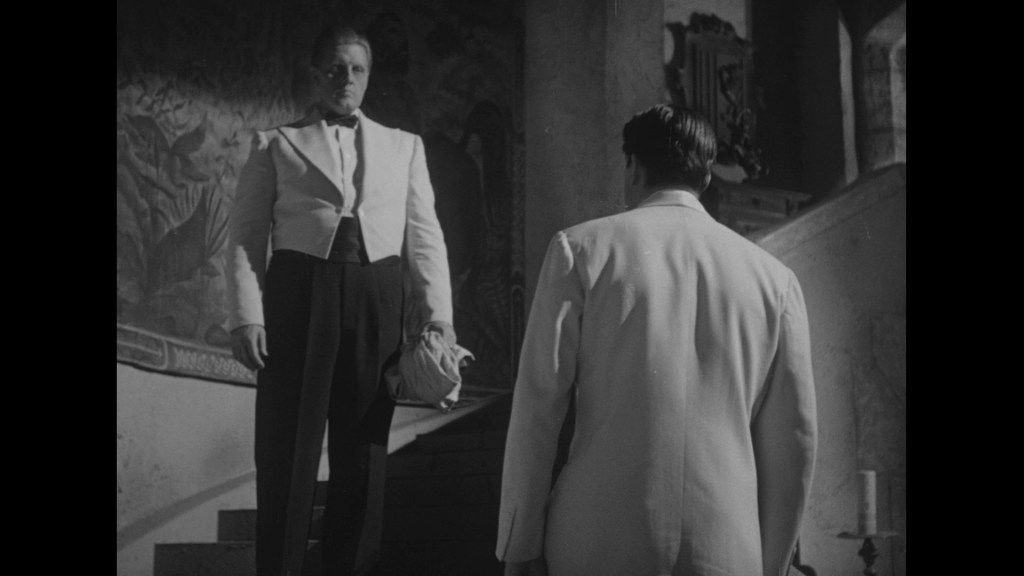 A dramatic black and white scene featuring two men dressed in formal attire. One man stands at the bottom of a staircase, looking up at the other man who stands at the top, partially turned away, creating a sense of tension.