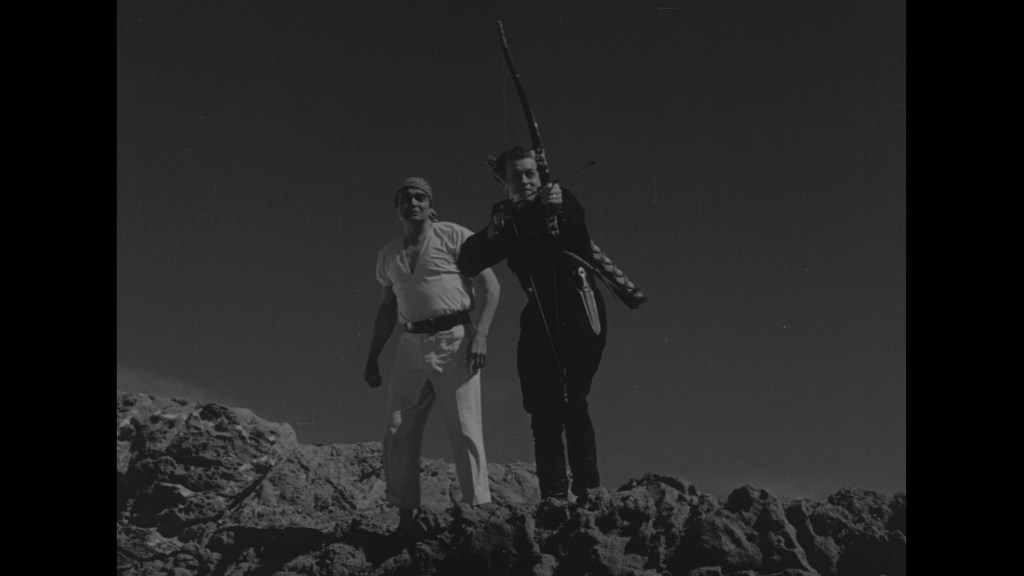 Two men standing on rocky terrain, one holding a rifle and the other observing.