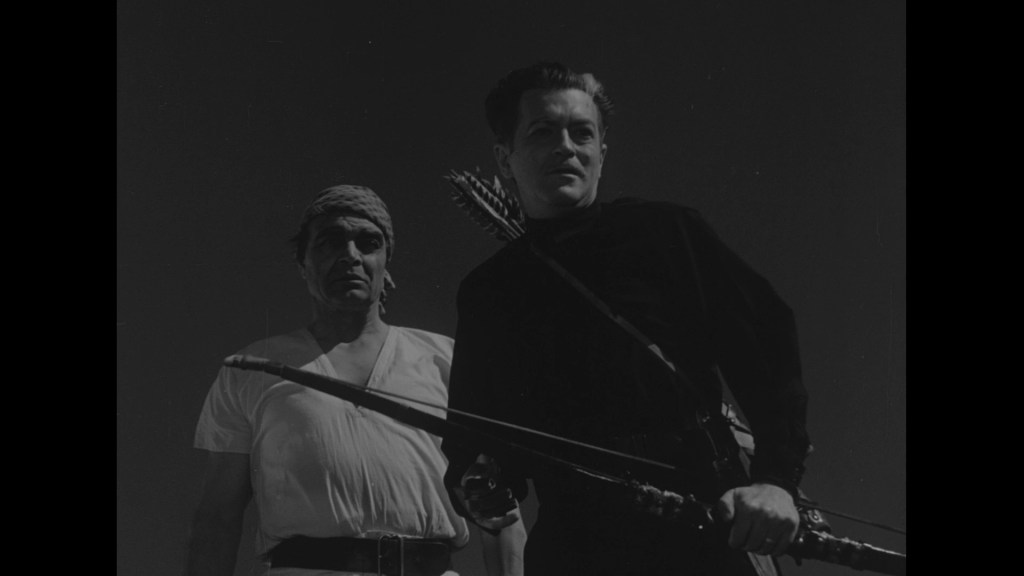 Two men standing in a dramatic pose, one wearing a black outfit holding a weapon, and the other in a white shirt with a head wrap.