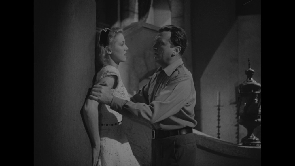 A man and a woman are in a close, dramatic encounter, with the man leaning towards the woman who is standing against a column. The scene is in black and white, featuring classic film styling.