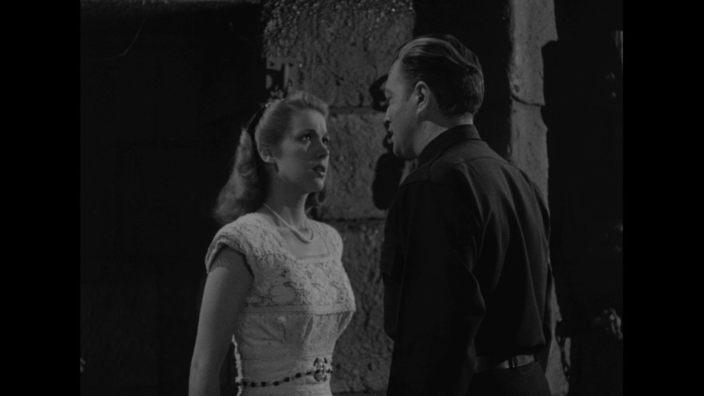 A dramatic black and white scene featuring a woman in a light-colored dress looking at a man in a dark uniform, framed in a textured stone wall.