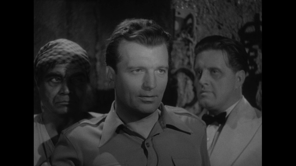 A scene featuring three men in a dramatic setting. The man in the foreground appears to be speaking intently, while the other two men, partially obscured in shadows, are listening closely.