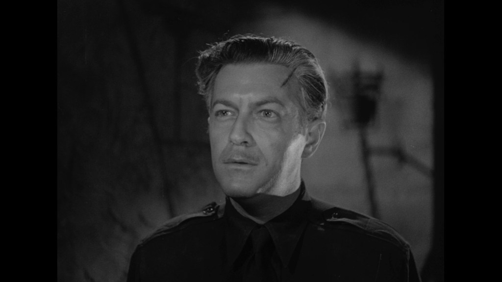 Close-up of a man with slicked-back hair and a serious expression, wearing a dark uniform, set against a shadowy backdrop.