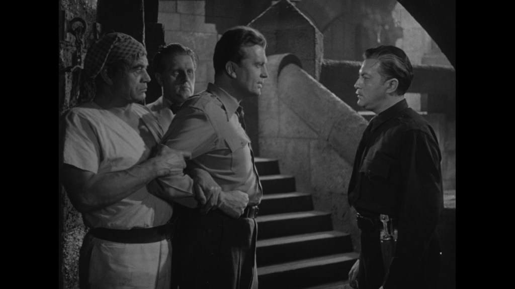 A black-and-white scene depicting four men in an underground setting, with one man in a police uniform confronting another who is restrained. The other two men observe the interaction.