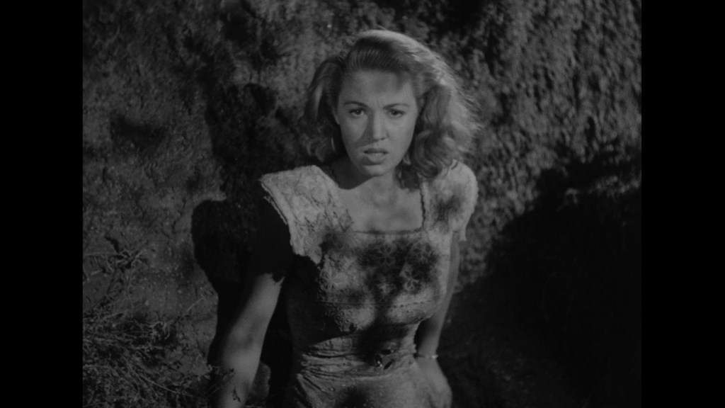 A woman with wavy hair wearing a textured dress looks directly at the camera with a concerned expression, standing in a dimly lit outdoor setting.