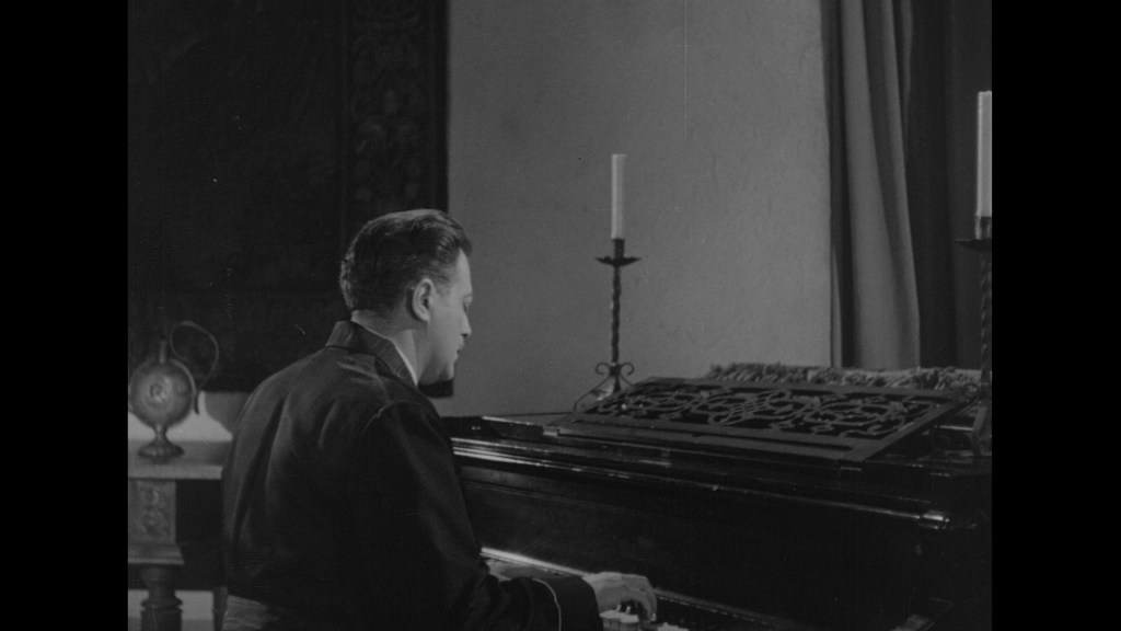A man seated at a piano, playing music, with candle holders on the piano and a decorative background.