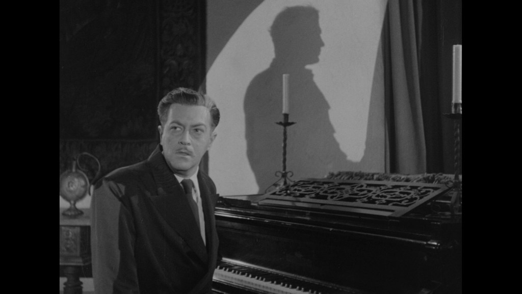 A man in formal attire stands near a piano, gazing thoughtfully to the side while a shadow is cast against a wall behind him.