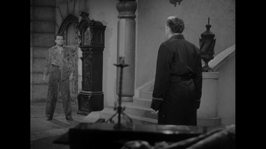 A tense scene featuring two men in a dimly lit room, one standing with his back to the camera wearing a robe, while the other stands slightly more forward, dressed in casual clothing.