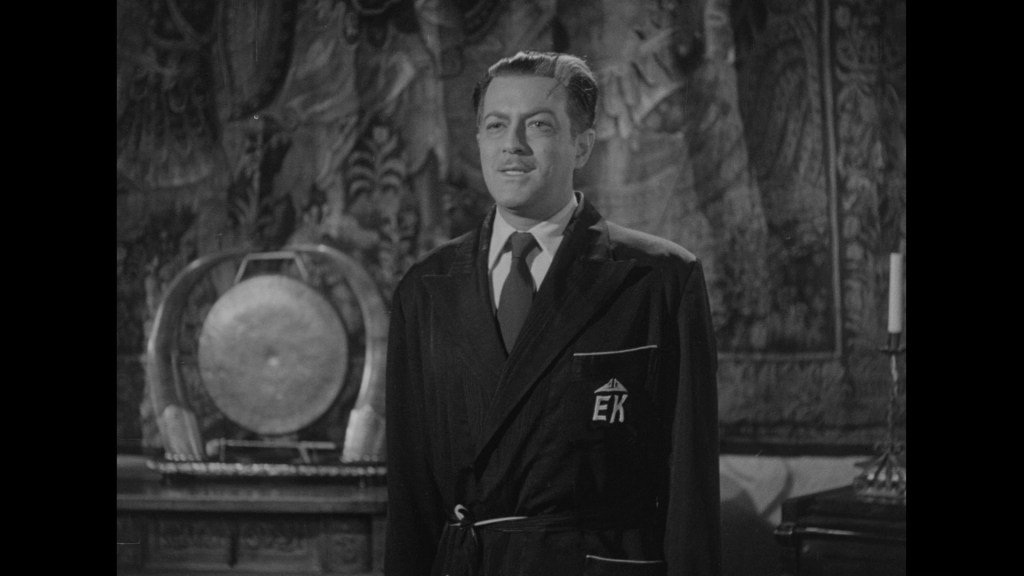A man in a black robe with 'EK' on the pocket stands in a decorated room with a tapestry, a gong, and a candle holder in the background.