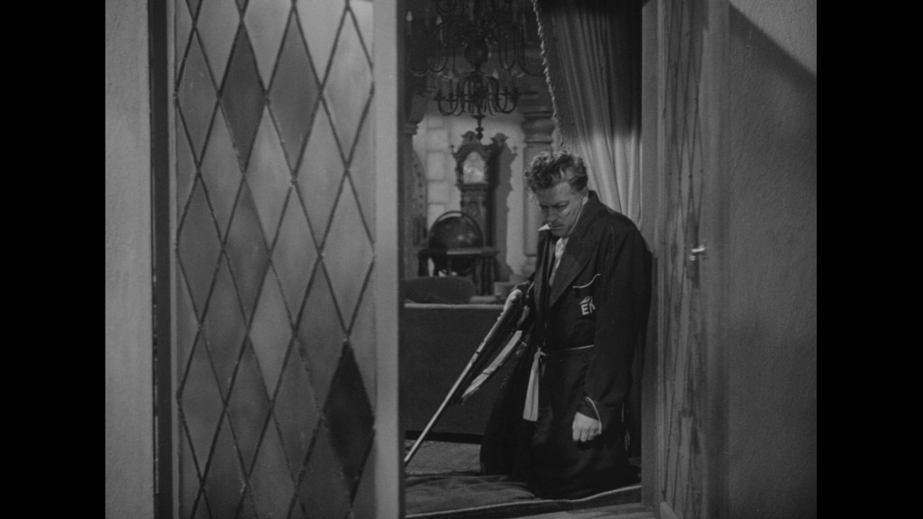 A man in a black robe kneels on the floor, holding a broom, in a dimly lit room with elegant decor.