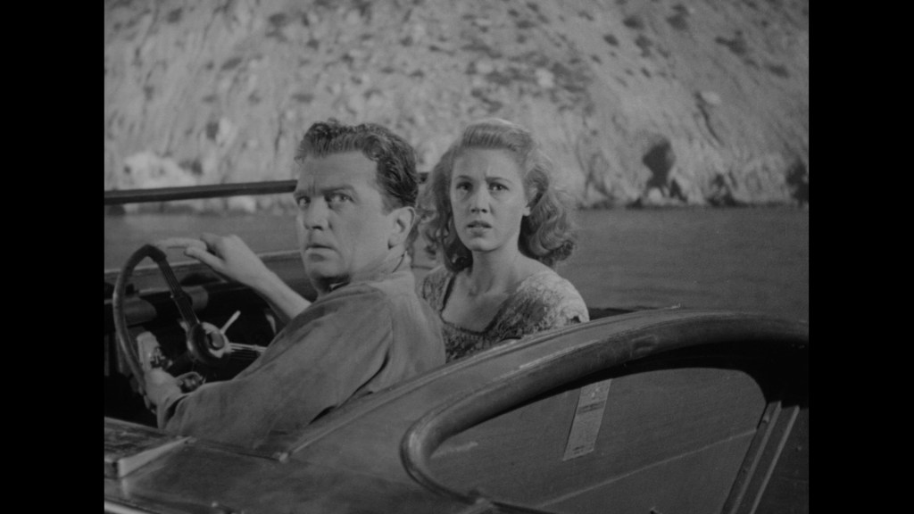 A man and a woman sit in a boat, looking alarmed as they face something off-screen. The scene is in black and white, with rocky cliffs in the background.