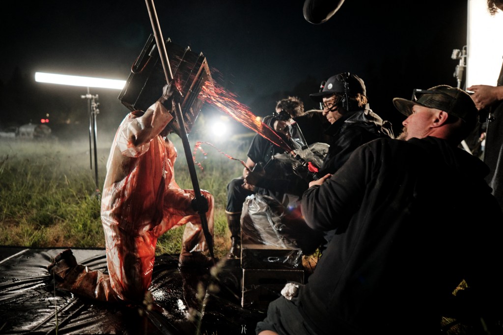 A film crew working on a night shoot, featuring a person in a blood-stained costume, simulating an intense scene with special effects. Lighting equipment and crew members are visible in the background.