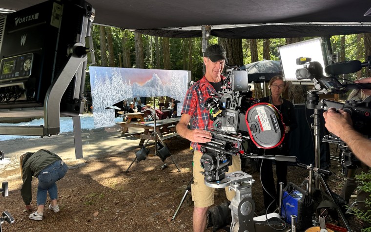 A film crew setting up equipment in a forested area, with a cinematographer operating a camera and a scenic backdrop of mountains and trees.