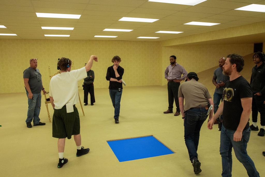 A group of people in a yellow-walled room with a blue square cutout in the floor, engaging in a discussion or demonstration.
